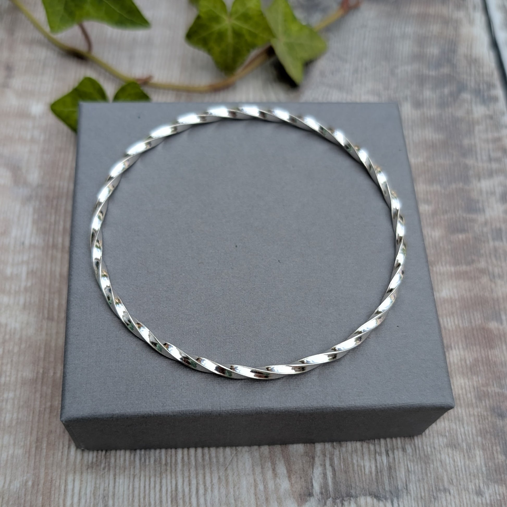 Silver Twisted Round Bangle handmade from recycled Sterling Silver 2mm square wire, shown resting on a grey jewellery box.