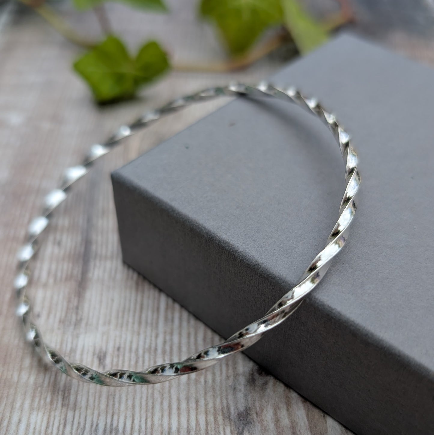 Silver Twisted Round Bangle handmade from recycled Sterling Silver 2mm square wire, displayed leaning against a grey jewellery box.