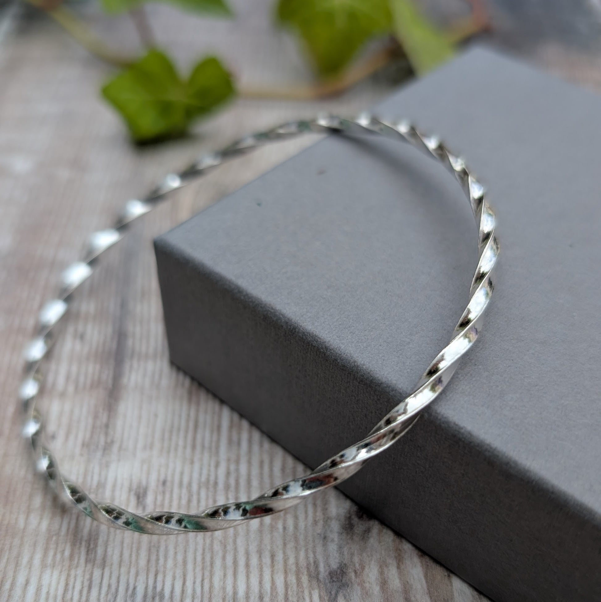 Silver Twisted Round Bangle handmade from recycled Sterling Silver 2mm square wire, displayed leaning against a grey jewellery box.