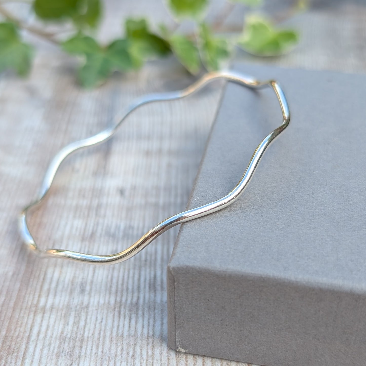 Silver Wavy Round Bangle handmade from recycled Sterling Silver displayed on the edge of a grey jewellery box, highlighting the smooth polished finish.