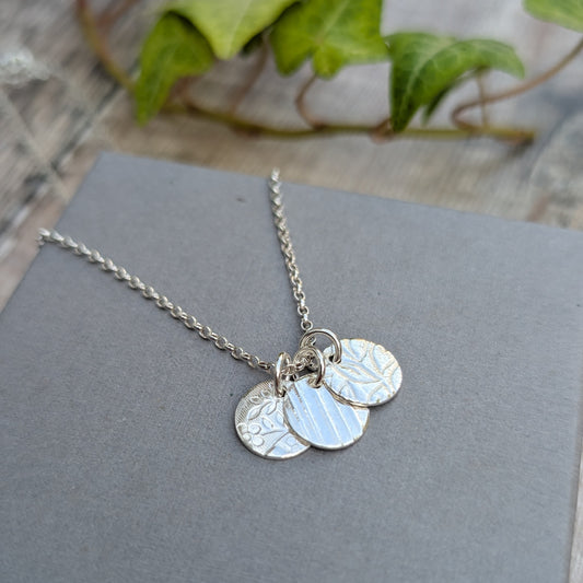 Silver wildflower trio of discs necklace handmade from recycled Sterling Silver, with three textured discs resting together on a grey background.