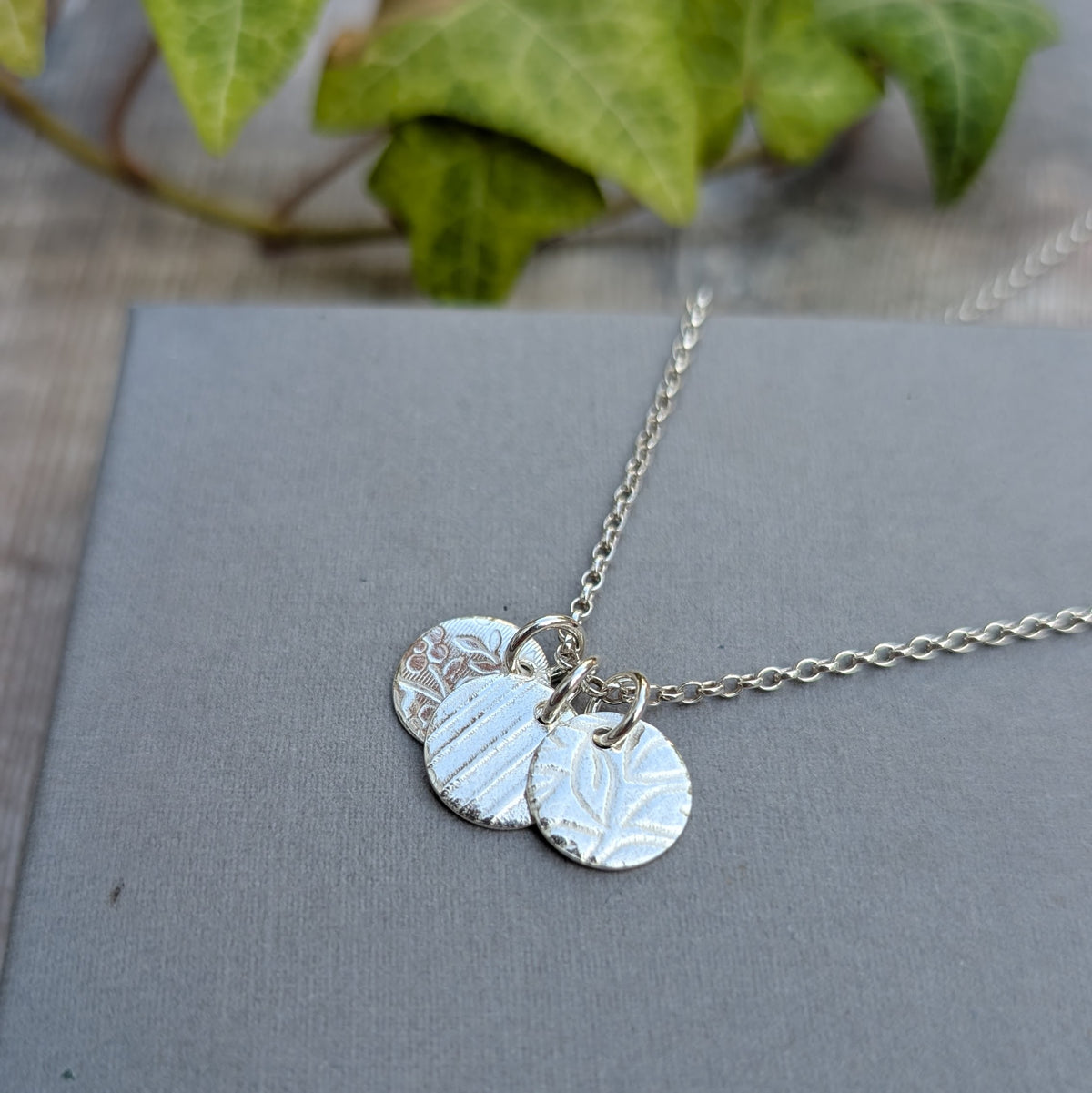 Close up of silver wildflower trio of discs necklace on a grey background, showing three textured recycled Sterling Silver discs on a chain.