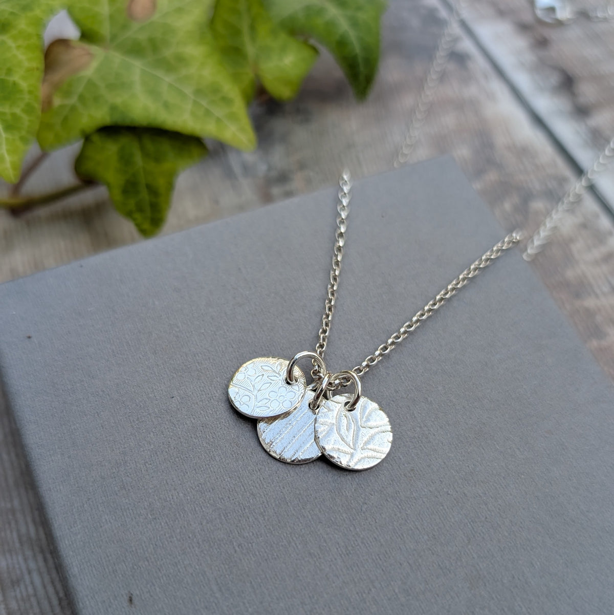 Silver wildflower trio of discs necklace with three overlapping patterned silver discs featuring floral, leaf and lined rolling mill textures.