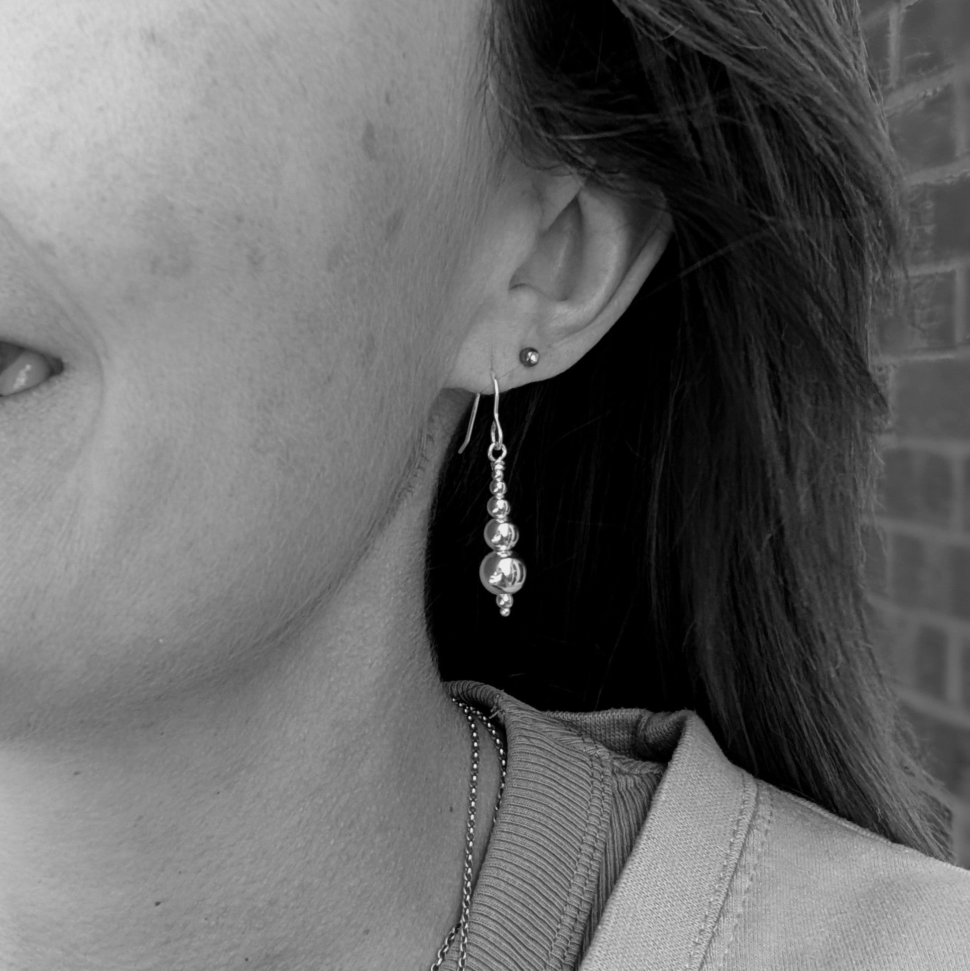 Black and white close-up of a person wearing a handmade sterling silver drop earring featuring polished round silver beads in descending sizes, with a second small stud earring visible in the upper ear.