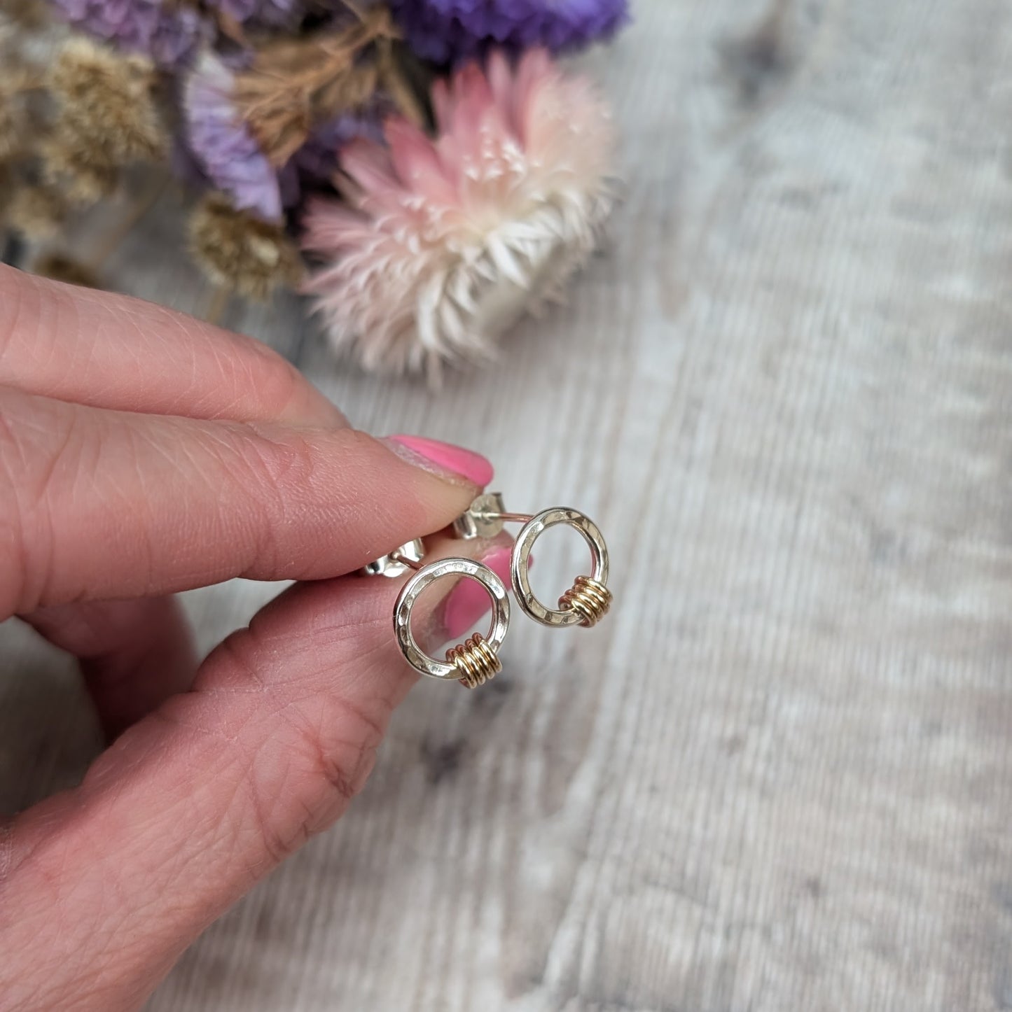 Silver circle stud earring with gold loops shown held between 2 fingers with dried flowers in the background. 