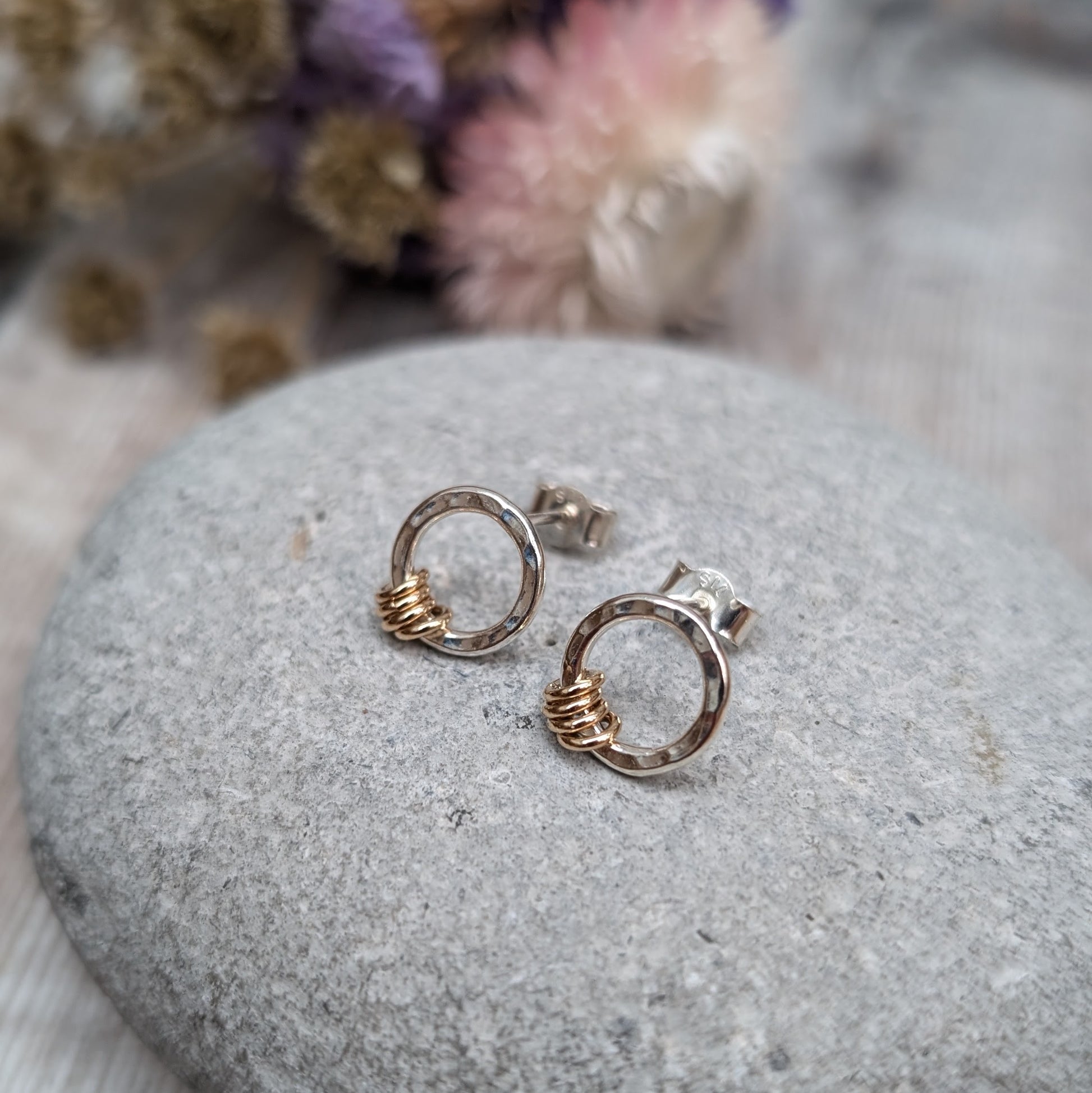 Silver circle stud earrings with gold loops shown displayed on a grey stone with dried flowers in the background. 