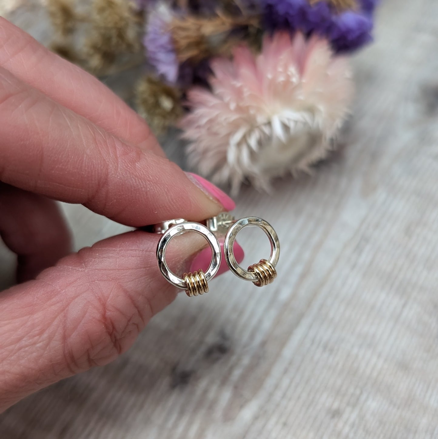 Silver circle stud earrings with gold loops shown held between 2 fingers with dried flowers in the background. 