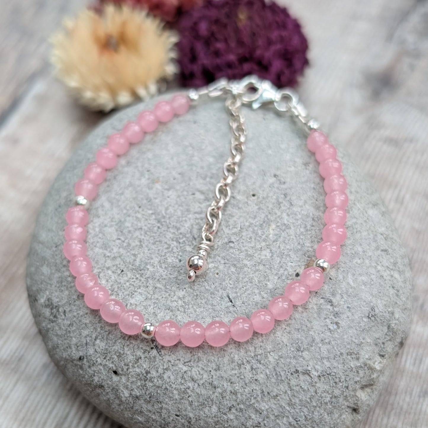 Rose quartz and sterling silver bead bracelet displayed on a smooth grey stone with dried flowers in the background, showing the clasp and extender chain.
