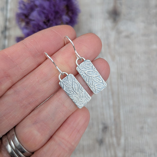 Close up of Silver small leaf pattern rectangle earrings in hand highlighting the pressed leaf pattern.