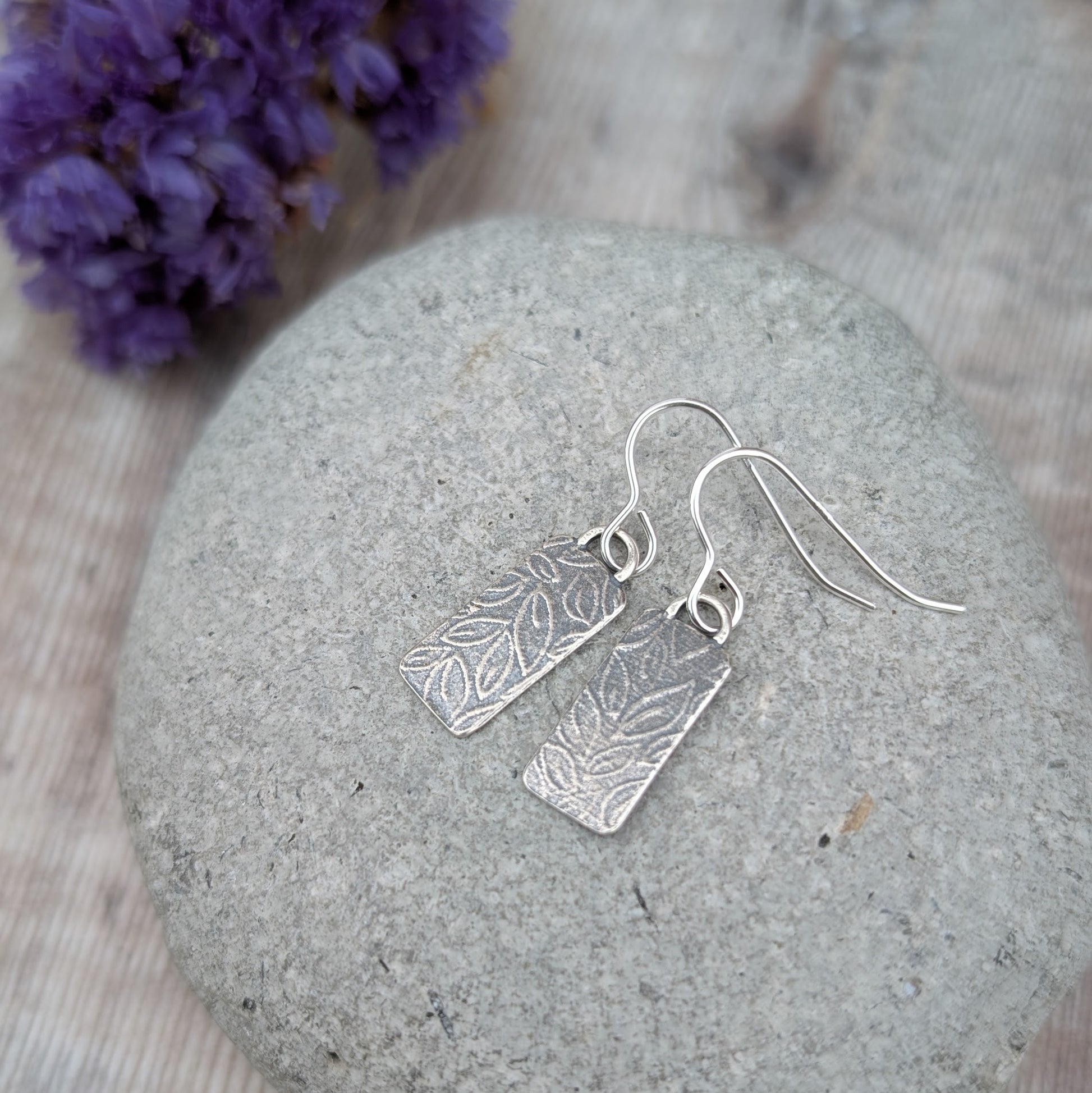 Silver small leaf pattern rectangle earrings resting on a smooth grey stone showing the matt oxidised finish.
