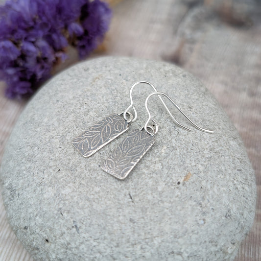 Silver small leaf pattern rectangle earrings displayed on a stone with purple flowers in the background.