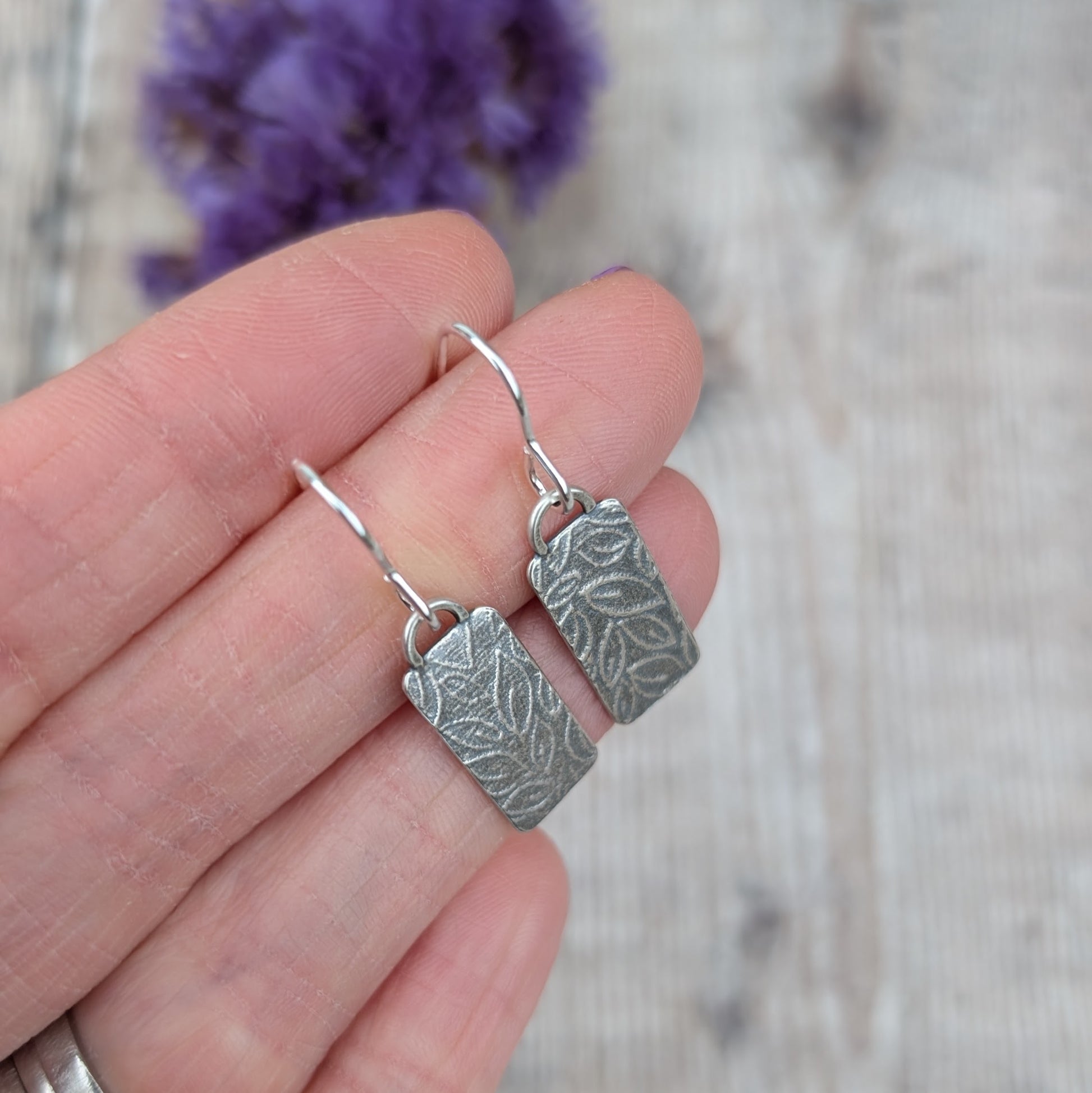 Silver small leaf pattern rectangle earrings held in a hand to show size and detail.