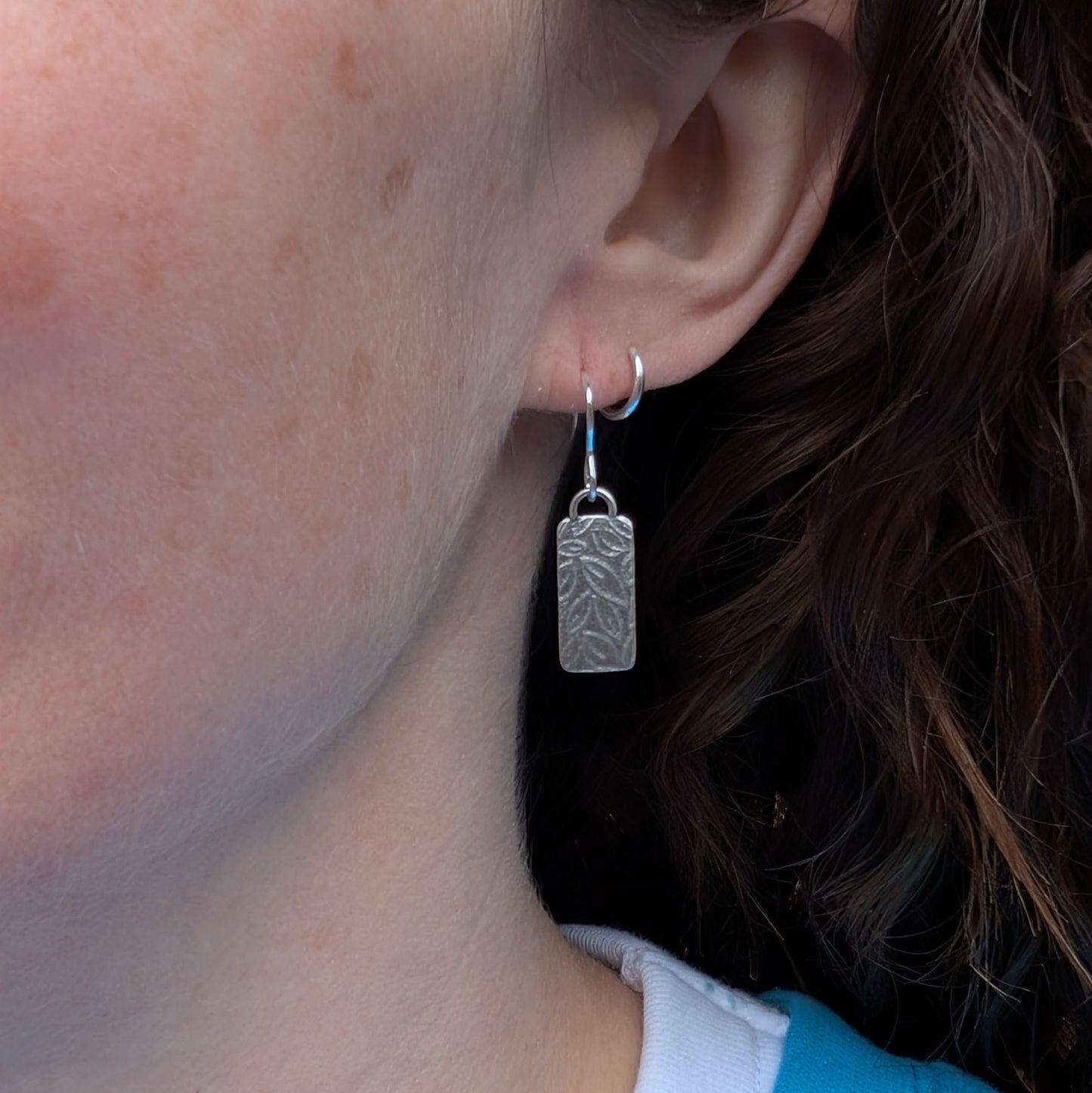 Silver small leaf pattern rectangle earrings worn on the ear showing how they hang.