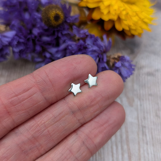 A close-up of a hand holding a pair of small silver star stud earrings, emphasizing their polished finish and minimalist look, with colorful flowers in the background.