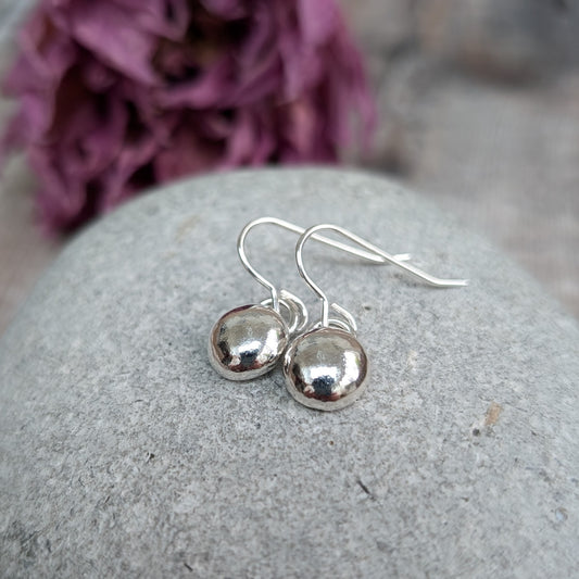 Pair of small silver dangle earrings with a smooth, polished pebble design, resting on a smooth stone. The reflective surface of the earrings contrasts with the matte stone, and a blurred purple flower is in the background.