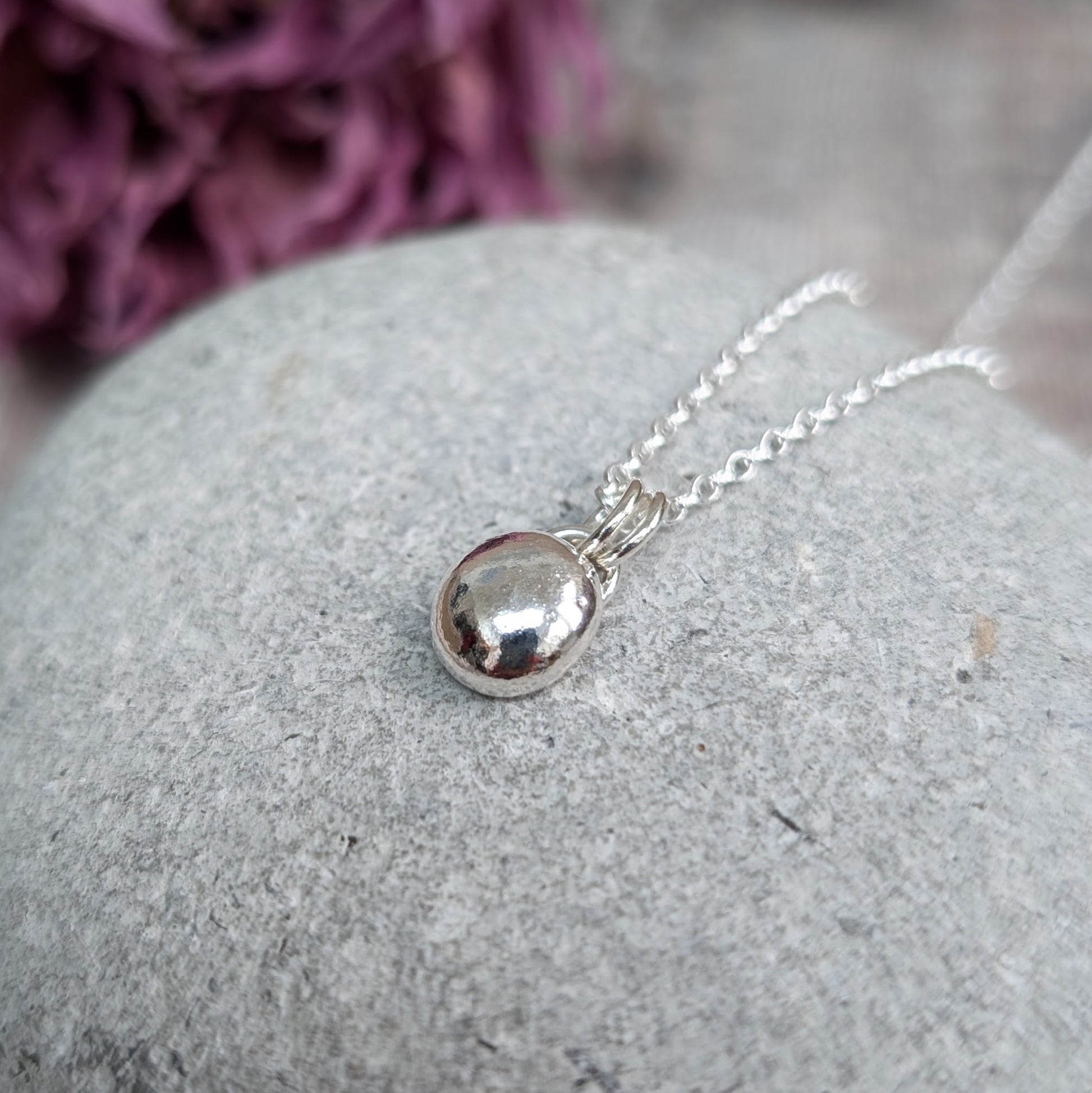 Close-up of a smooth pebble necklace with a shiny, rounded silver pendant on a delicate silver chain, resting on a light gray stone background.