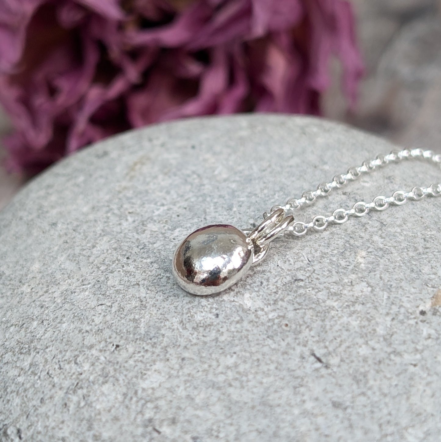 Close-up of a smooth pebble necklace with a shiny, rounded silver pendant on a delicate silver chain, resting on a light gray stone background.