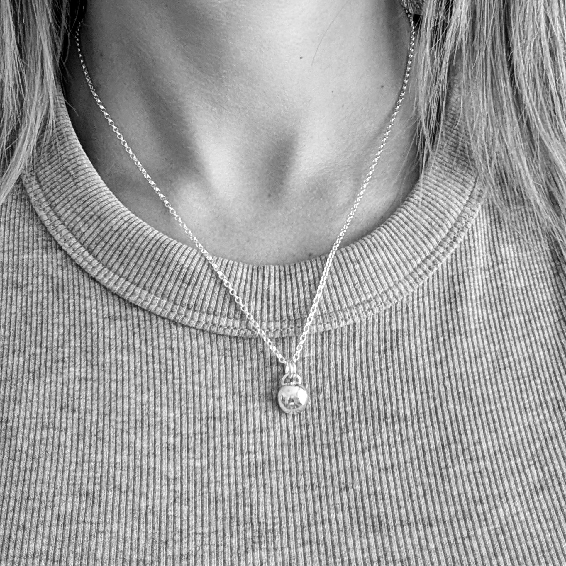 Black and white photo of a person wearing a silver necklace with a smooth pebble-shaped pendant, resting against a ribbed fabric top.