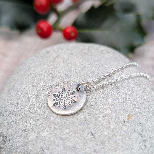 Silver disc pendant necklace featuring a single engraved snowflake design hand stamped onto the disc. Suspended from an 18 inch silver chain via a small open circle link.