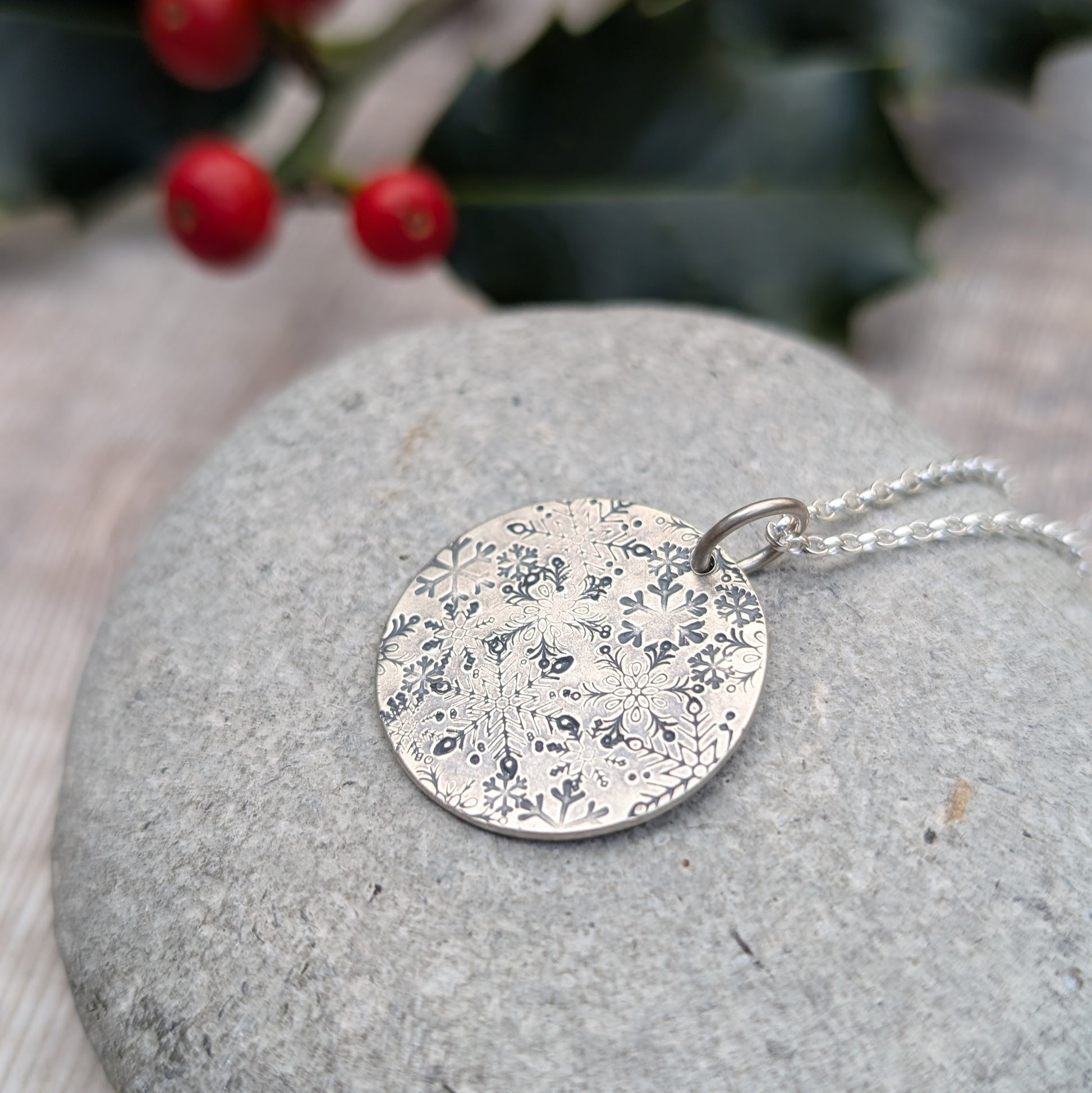 Pendant is a silver disc with intricate stamped snowflakes, some raised and some indented in texture. The snowflakes are randomly stamped and are all different in size and shape. Suspended from an 18 inch silver chain via a small open circle link.