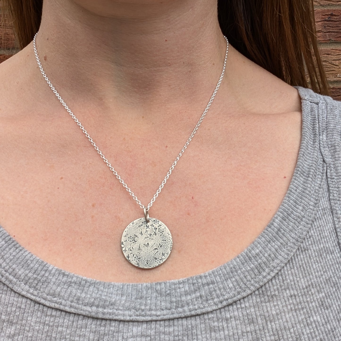 Pendant is a silver disc with intricate stamped snowflakes, some raised and some indented in texture. The snowflakes are randomly stamped and are all different in size and shape. Suspended from an 18 inch silver chain via a small open circle link.