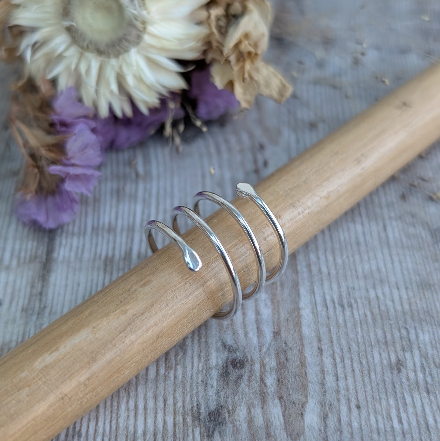 The silver spiral wraparound ring placed on a wooden ring mandrel, showing its shape and structure. The gentle curve and fluid design stand out against the natural wood grain, with dried flowers in the background.