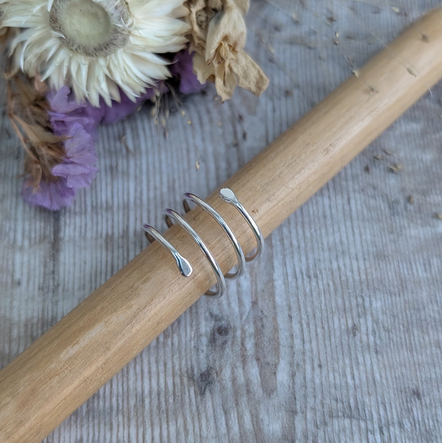 The silver spiral wraparound ring placed on a wooden ring mandrel, showing its shape and structure. The gentle curve and fluid design stand out against the natural wood grain, with dried flowers in the background.