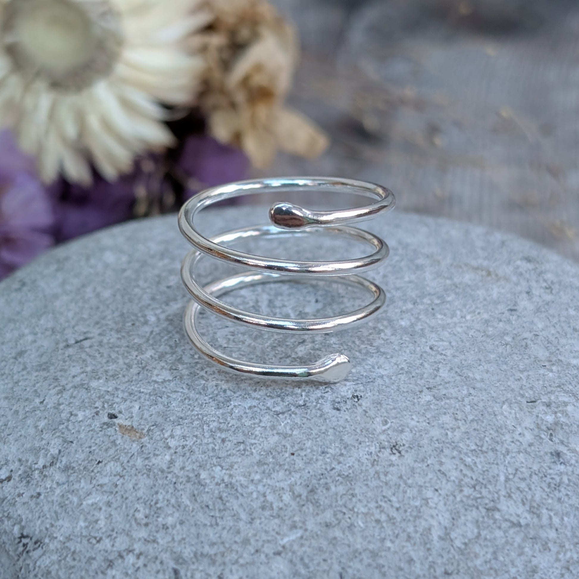 A handmade sterling silver spiral wraparound ring resting on a smooth grey stone. The ring features a flowing, coiled design with tapered ends, reflecting light. Dried flowers in the background add a soft, natural touch.