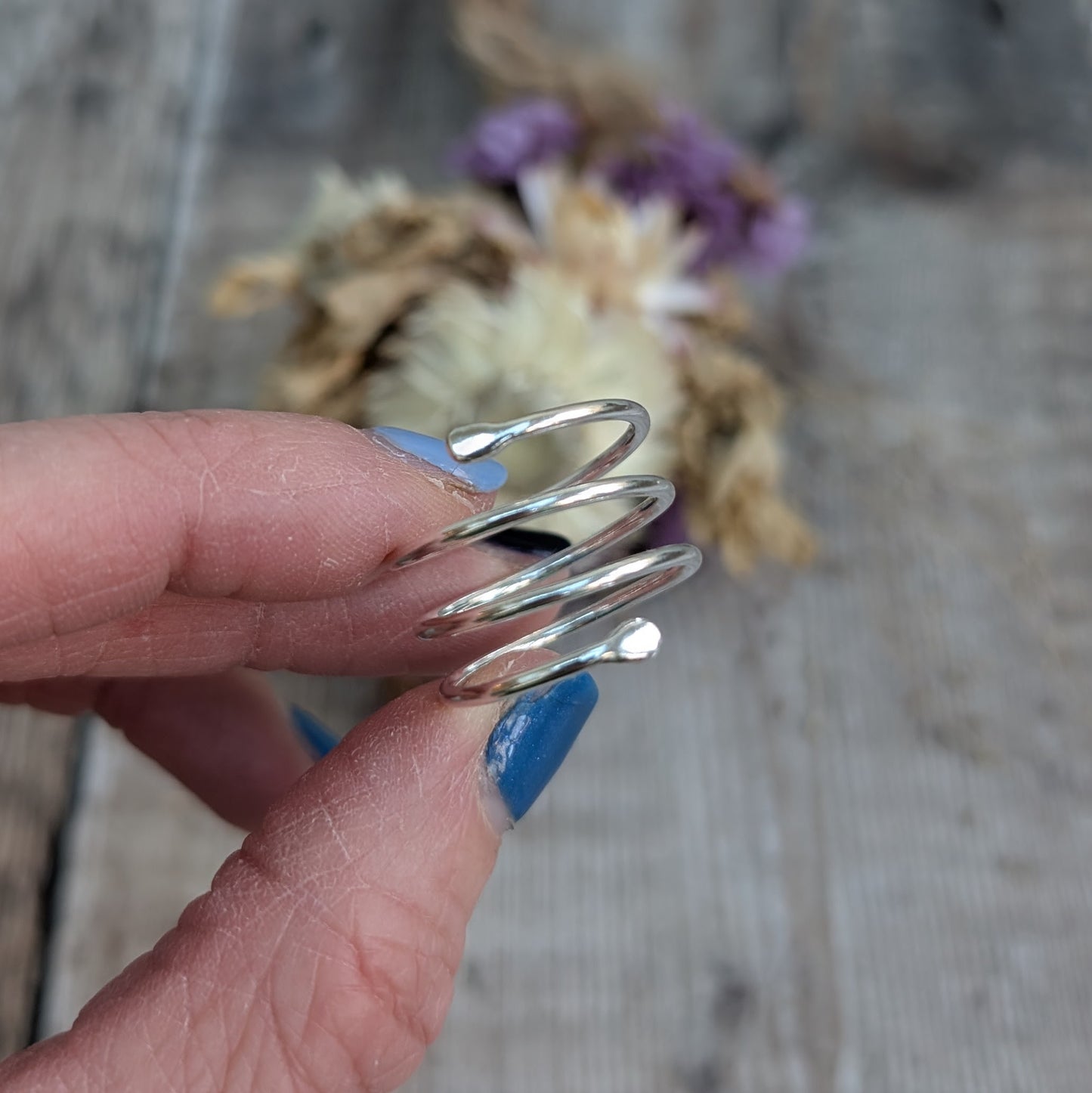 The silver spiral ring held between fingers, showing its open-ended, wraparound design. The polished surface catches the light, and a blurred background of dried flowers provides a warm contrast.
