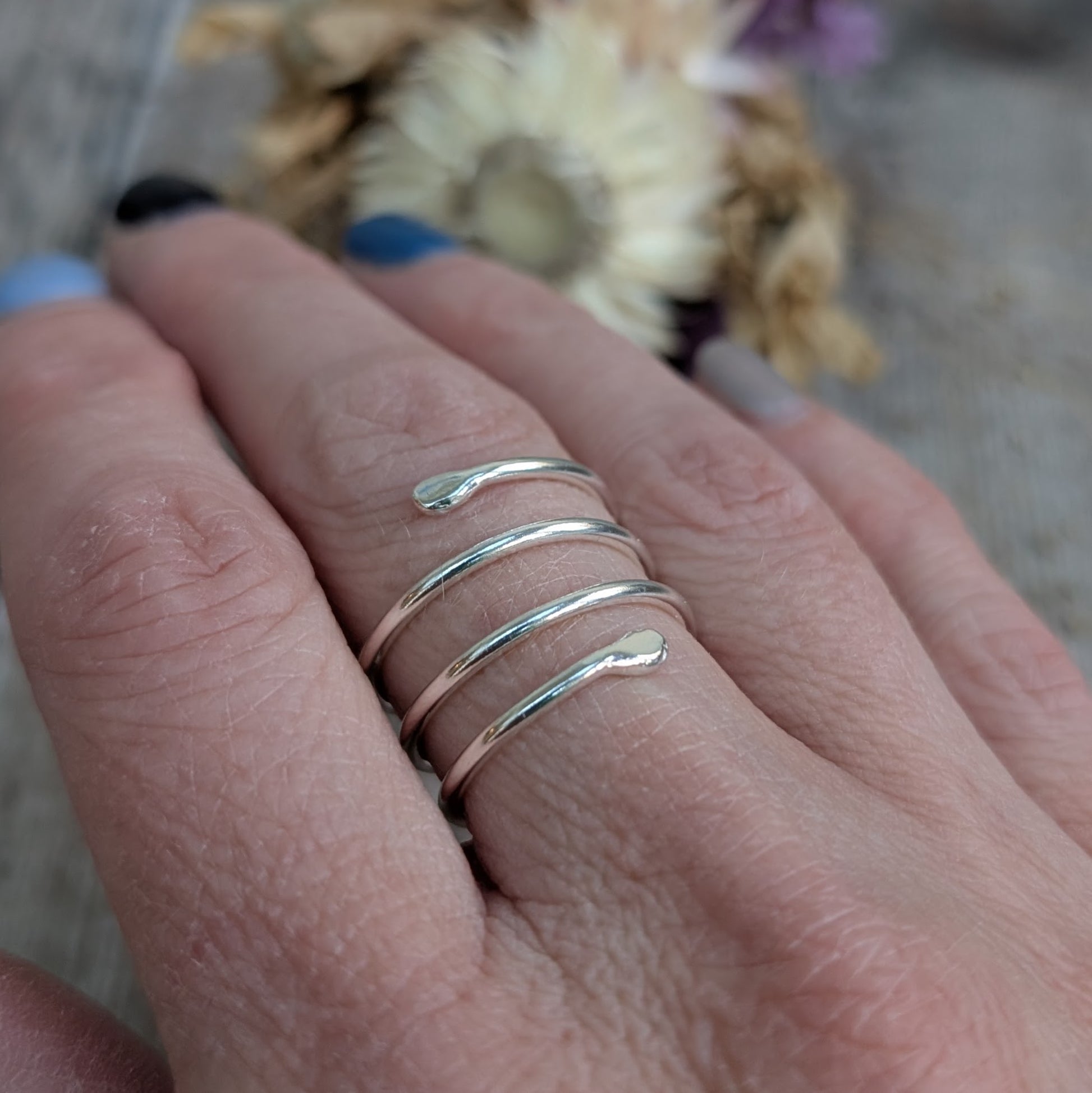 The silver spiral ring worn on a finger, wrapping around elegantly. The open-ended design allows for slight adjustability, and the smooth, rounded tips create a comfortable fit. The wearer’s nails are painted in soft shades of blue and black.