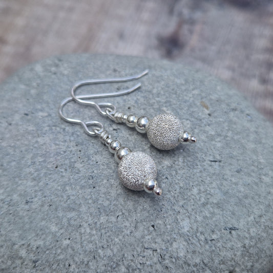 A close-up of sterling silver earrings with stardust beads and polished silver details, resting on a smooth grey stone. The shimmering texture of the stardust finish contrasts beautifully with the sleek silver components.