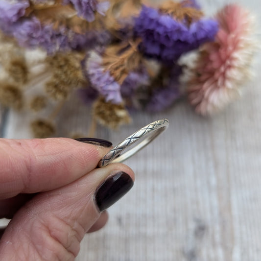 Close-up of a hand holding a delicate silver ring with a crosshatch pattern called "Starlight," set against a soft background of dried flowers in purple and pink hues.