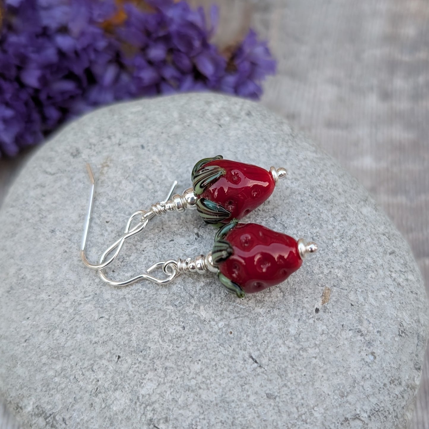 A pair of handmade earrings featuring vibrant strawberry-shaped lampwork glass beads with green leaves, dangling from sterling silver hooks. The earrings are displayed on a smooth grey stone with a purple floral background.