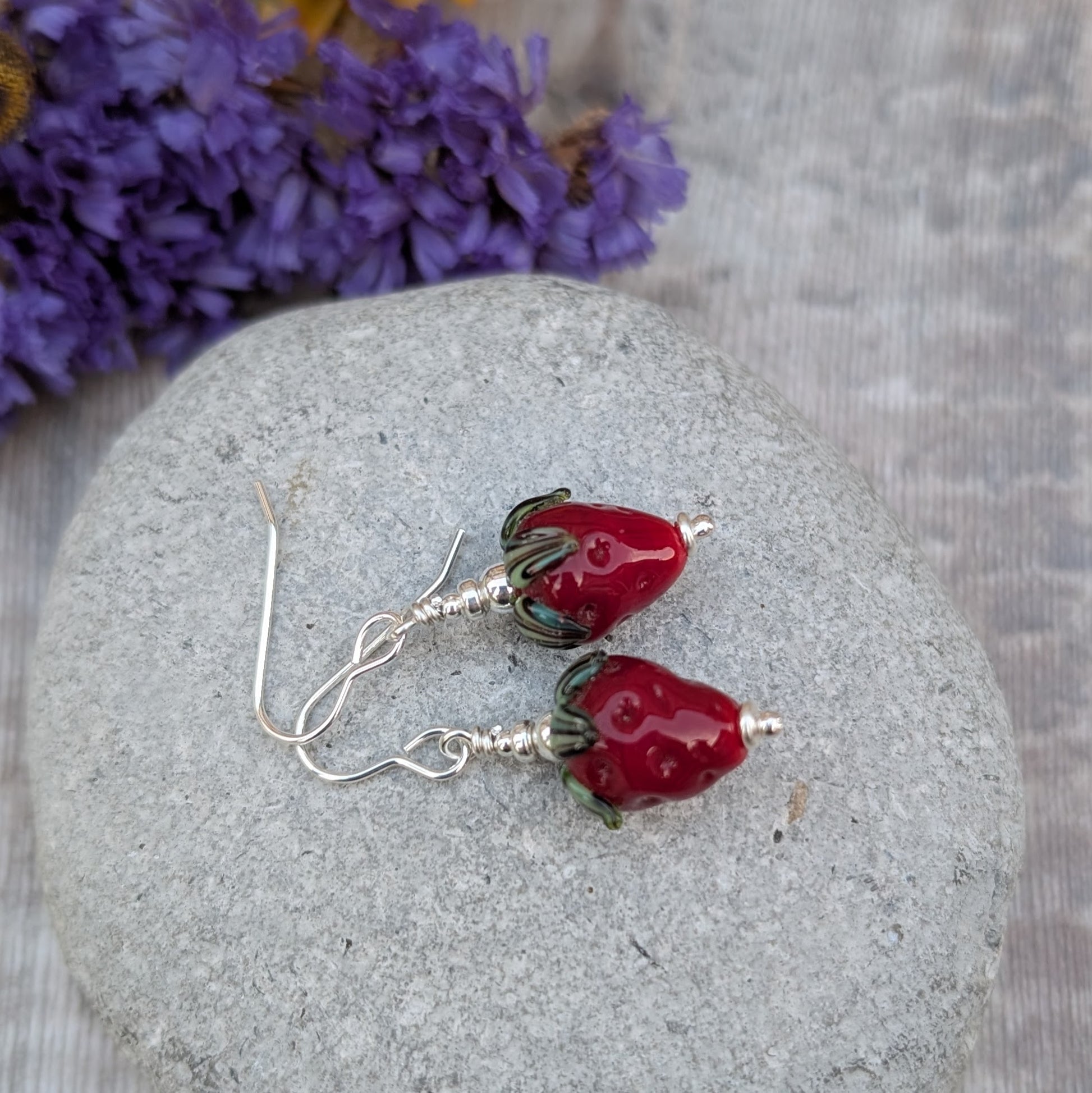 A pair of handmade earrings featuring vibrant strawberry-shaped lampwork glass beads with green leaves, dangling from sterling silver hooks. The earrings are displayed on a smooth grey stone with a purple floral background.