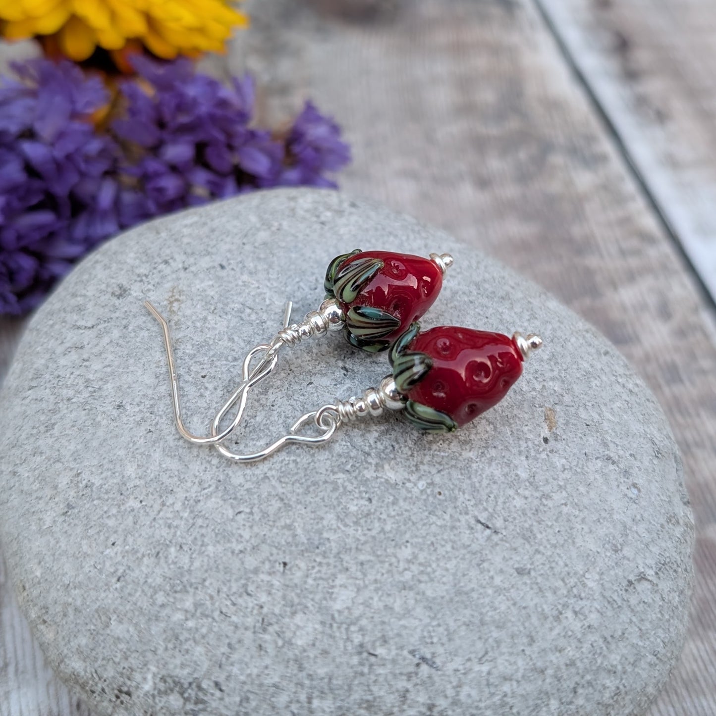 A pair of handmade earrings featuring vibrant strawberry-shaped lampwork glass beads with green leaves, dangling from sterling silver hooks. The earrings are displayed on a smooth grey stone with a purple floral background.