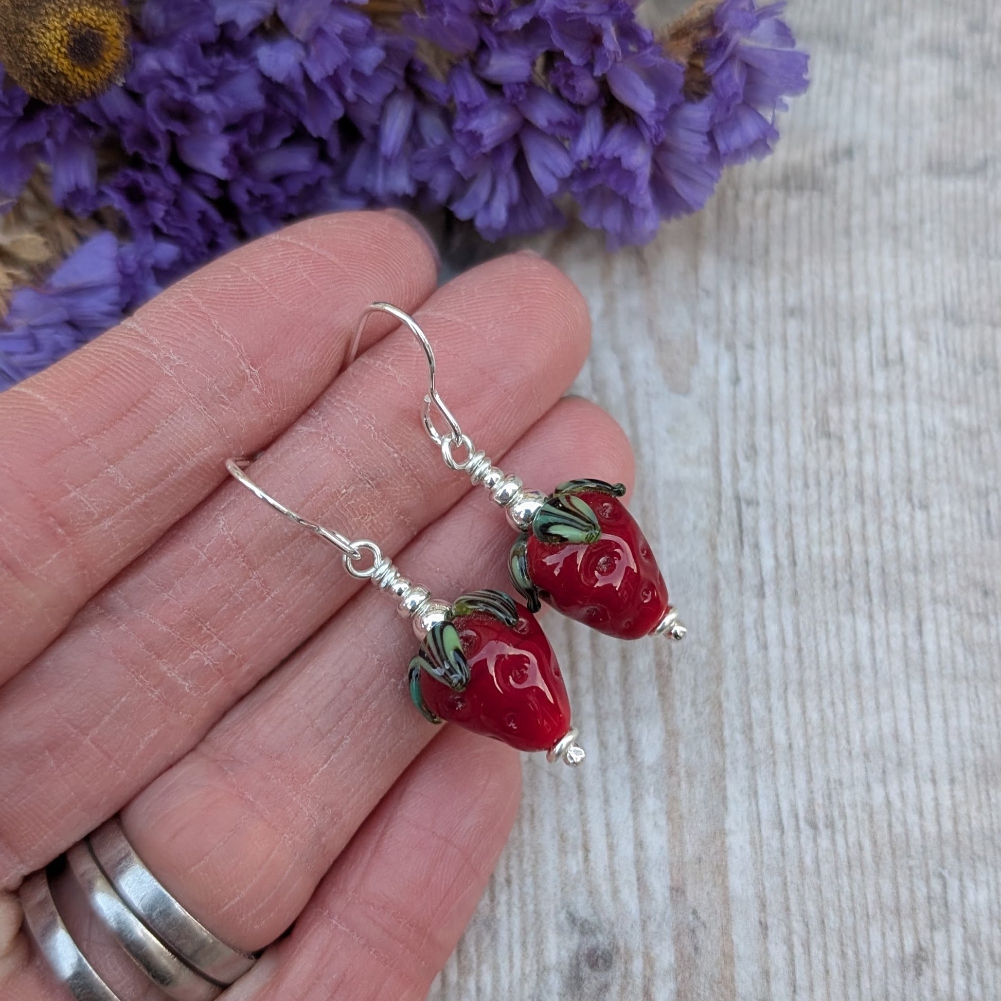 A close-up of a hand holding a pair of handmade strawberry lampwork glass bead earrings, showcasing their detailed design and sterling silver hooks. The background includes blurred purple flowers.