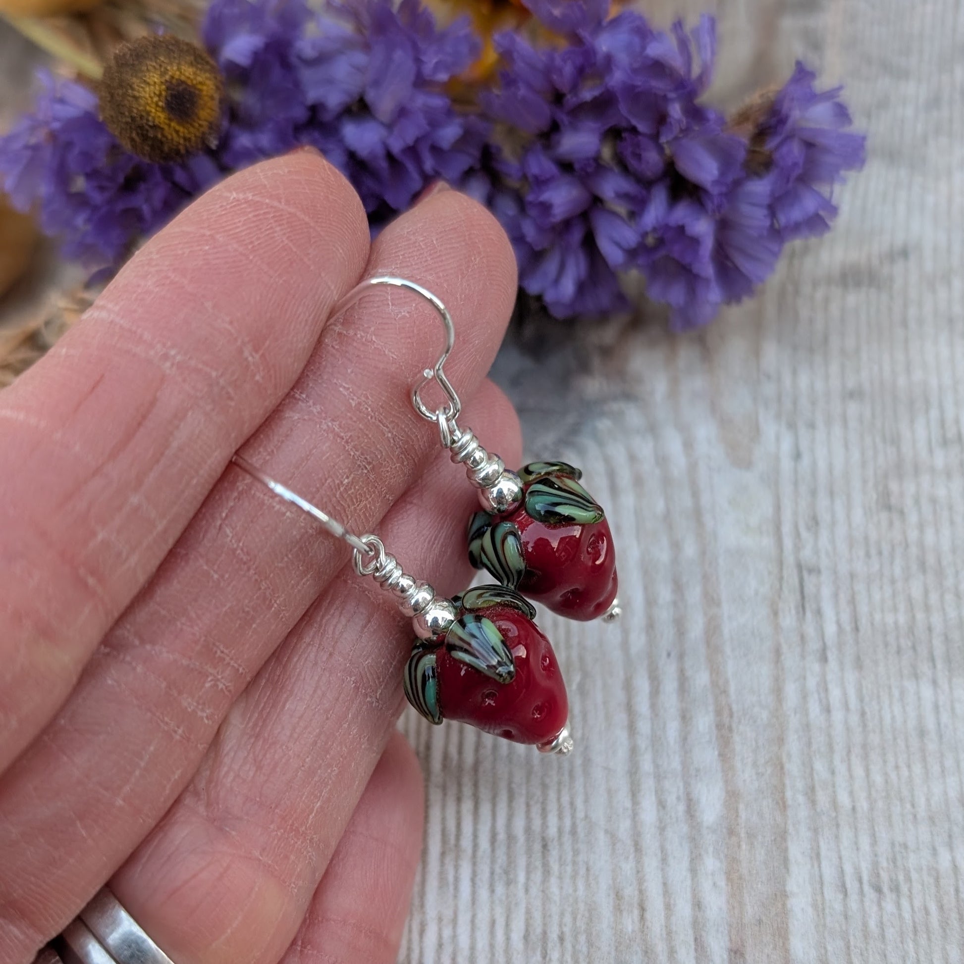 A close-up of a hand holding a pair of handmade strawberry lampwork glass bead earrings, showcasing their detailed design and sterling silver hooks. The background includes blurred purple flowers.