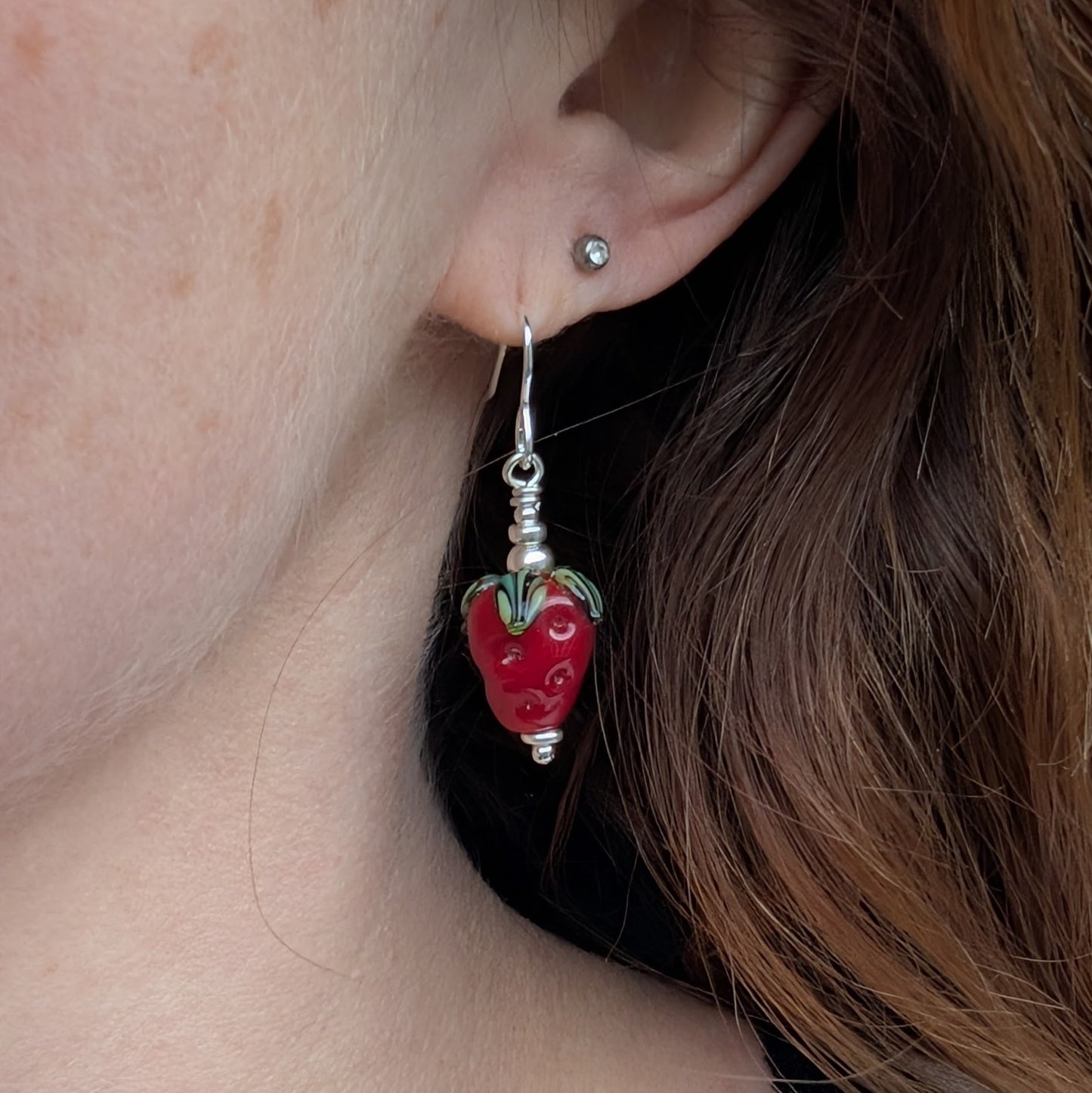 A single strawberry-shaped lampwork glass bead earring being worn, highlighting its size and vibrant red and green details. The sterling silver hook is visible, complementing the playful design.