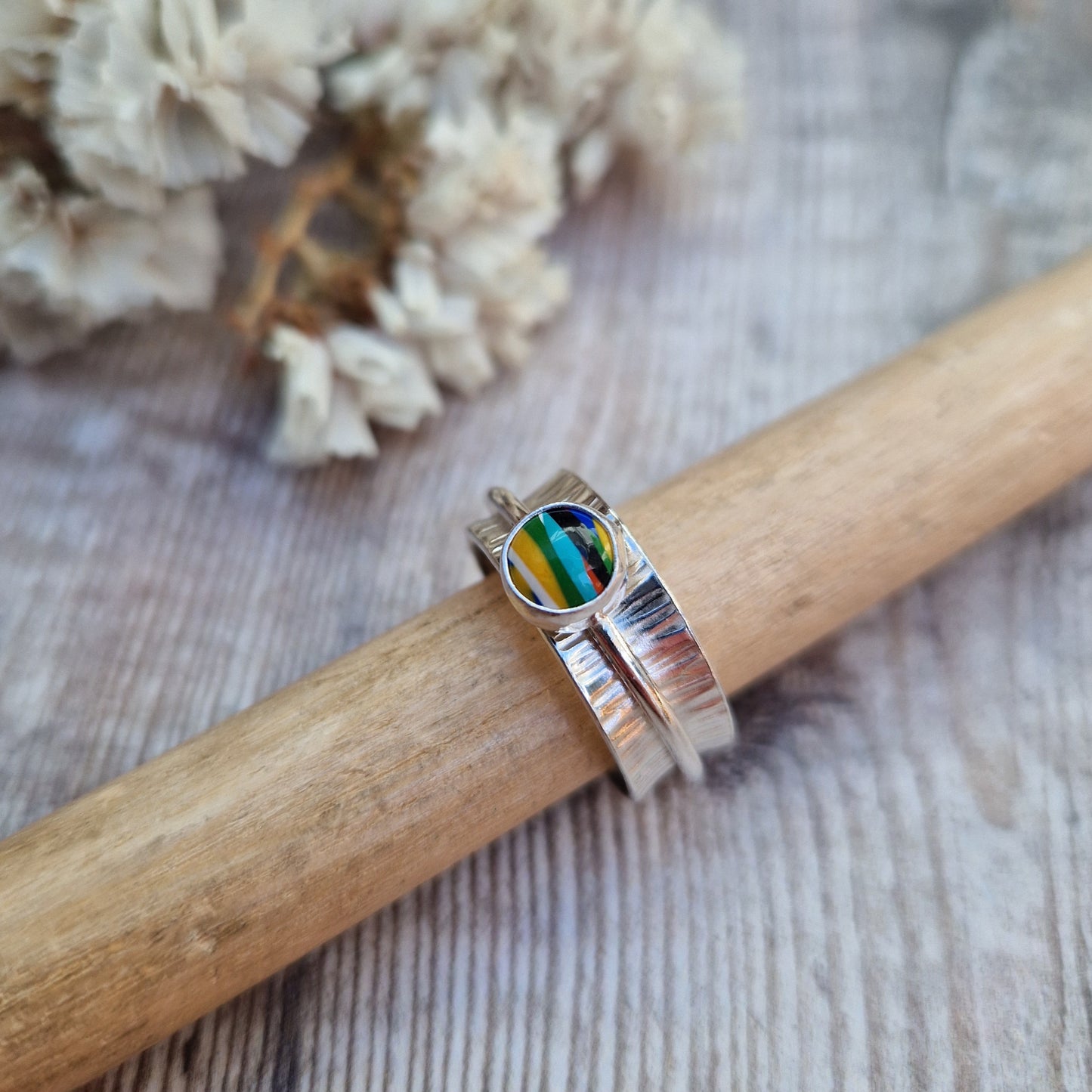 Approx. 8mm wide textured silver band with additional 2mm wide silver band around the middle that spins independently. Mounted in a silver bezel on top of the 2mm wide band is a round surfite stone, approx. 6mm diameter. Diagonal striped colours in the surfite are yellow, blue, black, aqua blue, green, yellow and white. 