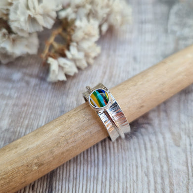 Approx. 8mm wide textured silver band with additional 2mm wide silver band around the middle that spins independently. Mounted in a silver bezel on top of the 2mm wide band is a round surfite stone, approx. 6mm diameter. Diagonal striped colours in the surfite are yellow, blue, black, aqua blue, green, yellow and white. 