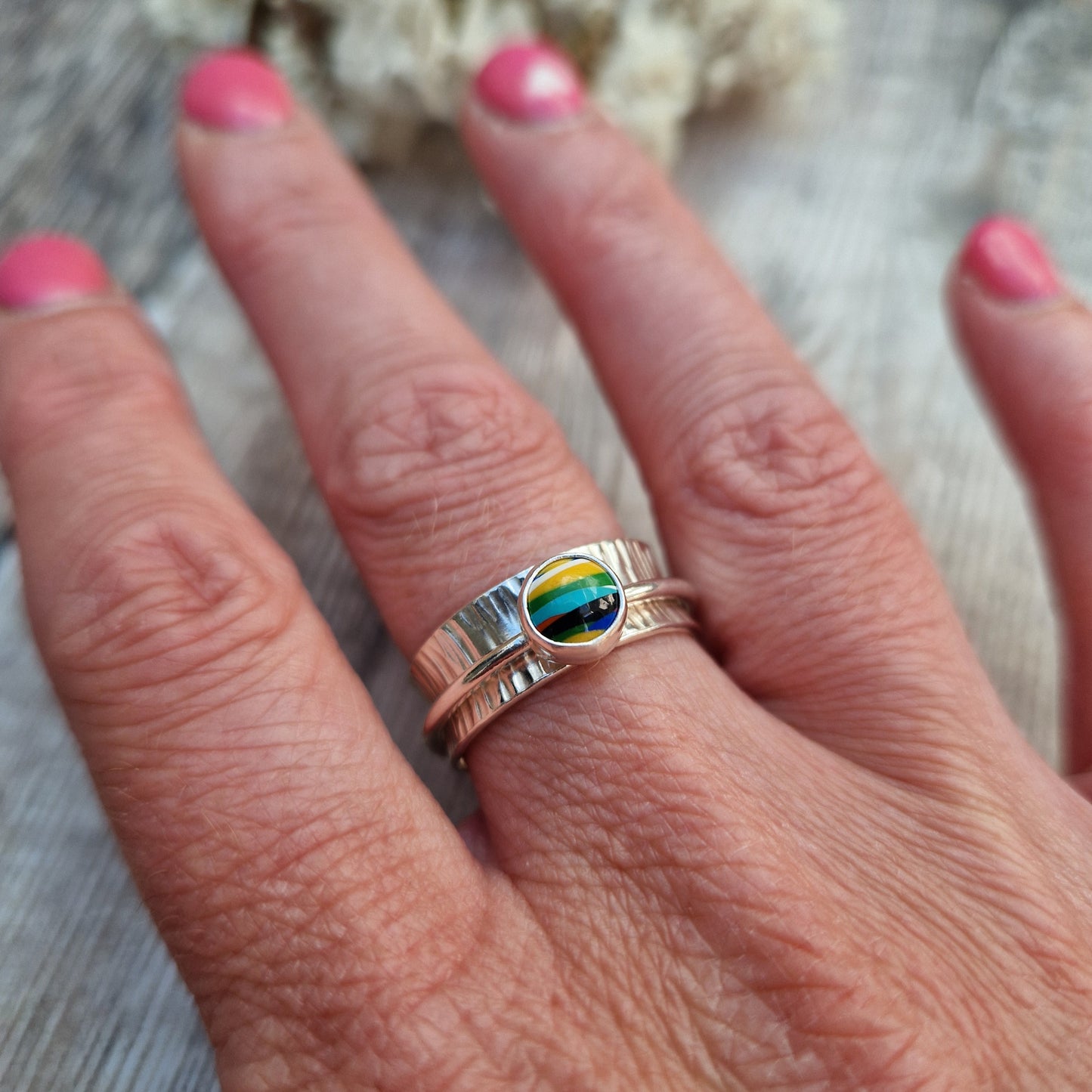 Approx. 8mm wide textured silver band with additional 2mm wide silver band around the middle that spins independently. Mounted in a silver bezel on top of the 2mm wide band is a round surfite stone, approx. 6mm diameter. Diagonal striped colours in the surfite are yellow, blue, black, aqua blue, green, yellow and white. 