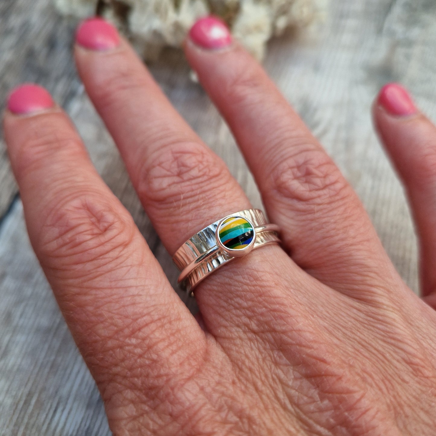 Approx. 8mm wide textured silver band with additional 2mm wide silver band around the middle that spins independently. Mounted in a silver bezel on top of the 2mm wide band is a round surfite stone, approx. 6mm diameter. Diagonal striped colours in the surfite are yellow, blue, black, aqua blue, green, yellow and white. 