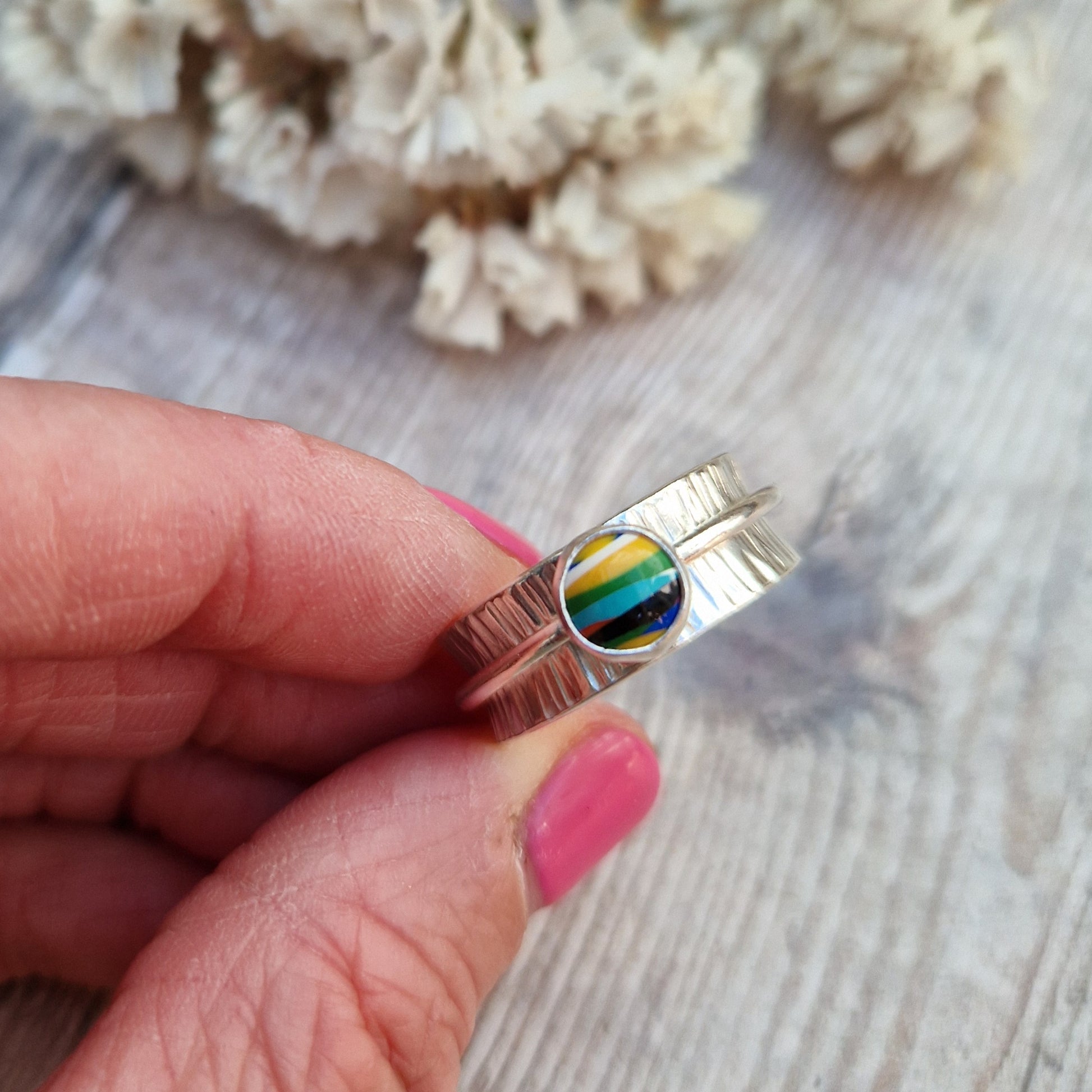 Approx. 8mm wide textured silver band with additional 2mm wide silver band around the middle that spins independently. Mounted in a silver bezel on top of the 2mm wide band is a round surfite stone, approx. 6mm diameter. Diagonal striped colours in the surfite are yellow, blue, black, aqua blue, green, yellow and white. 