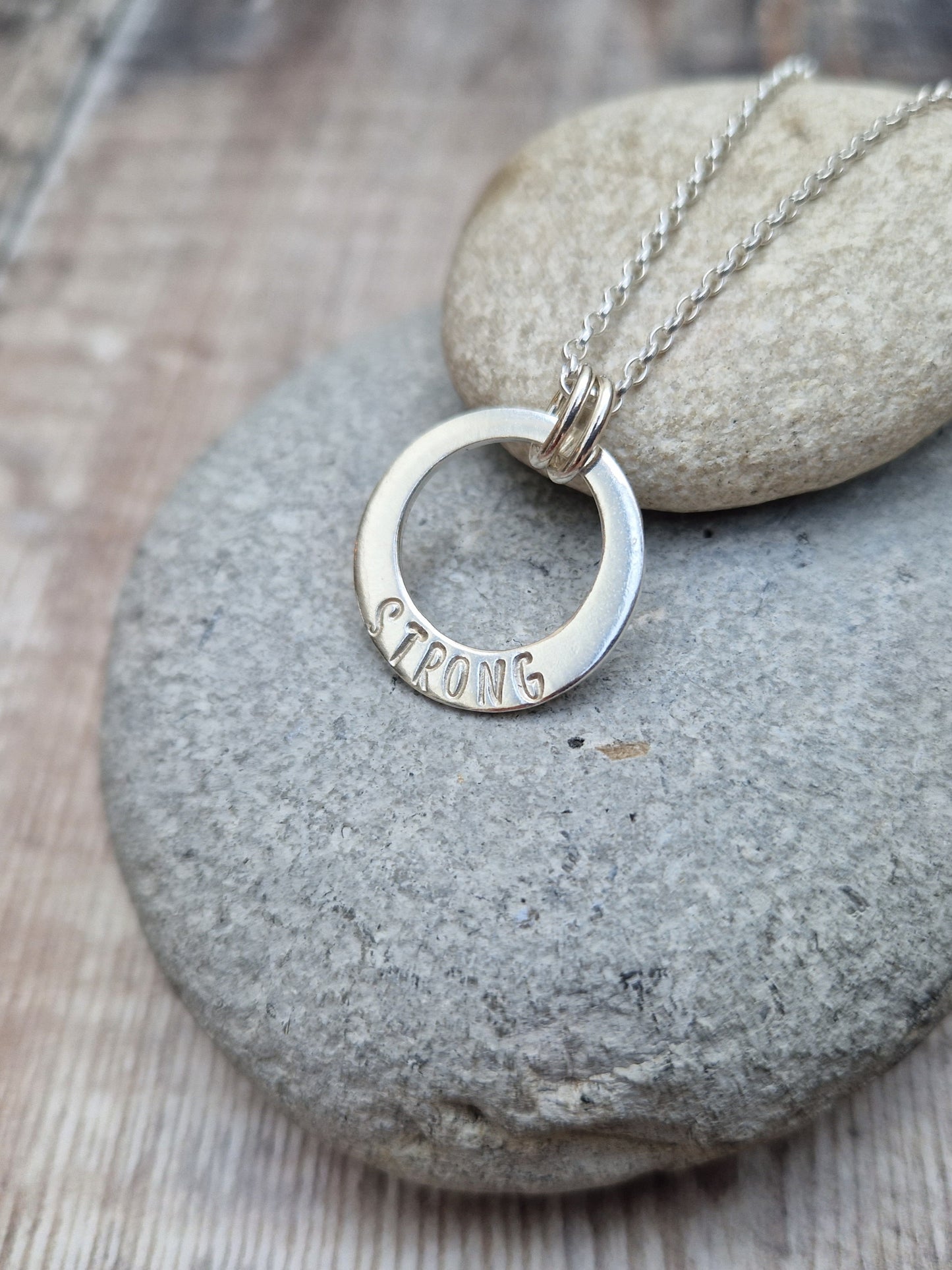 Sterling Silver ‘STRONG’ necklace. Silver disc with offset hole in centre, looks like a thick circle. Attached via two small silver hoops to silver chain. On thicker part of disc, inscription wording hammered into the silver reads STRONG in capitals. Disc measures approximately 20 mm in diameter. Chain length 16, 18 or 20 inches.