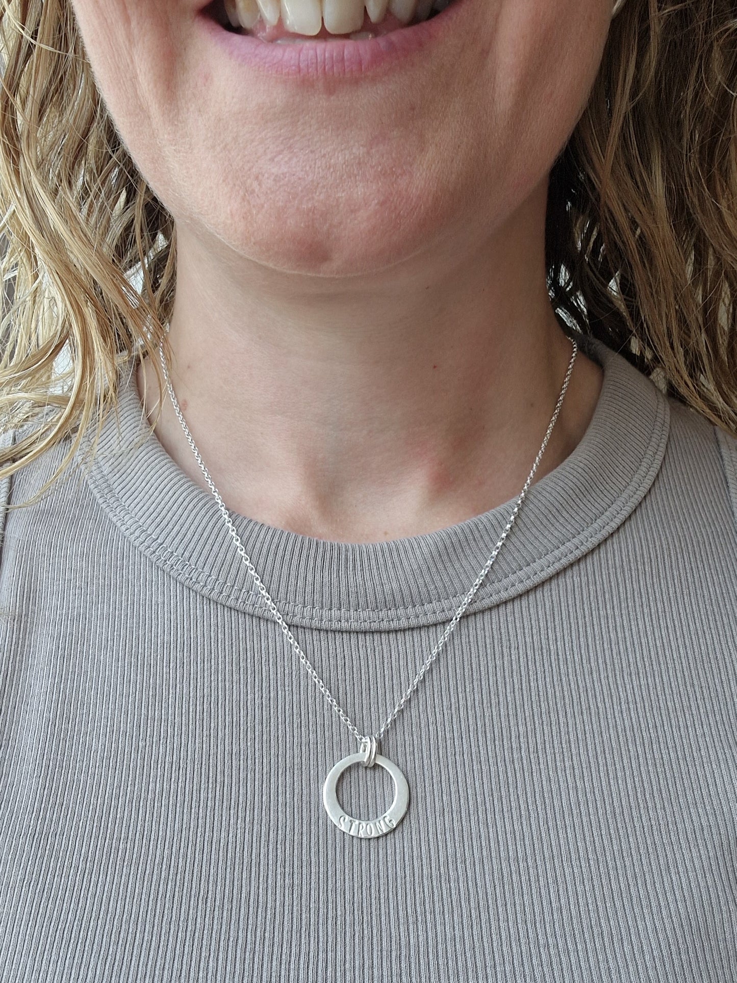 Sterling Silver ‘STRONG’ necklace. Silver disc with offset hole in centre, looks like a thick circle. Attached via two small silver hoops to silver chain. On thicker part of disc, inscription wording hammered into the silver reads STRONG in capitals. Disc measures approximately 20 mm in diameter. Chain length 16, 18 or 20 inches.