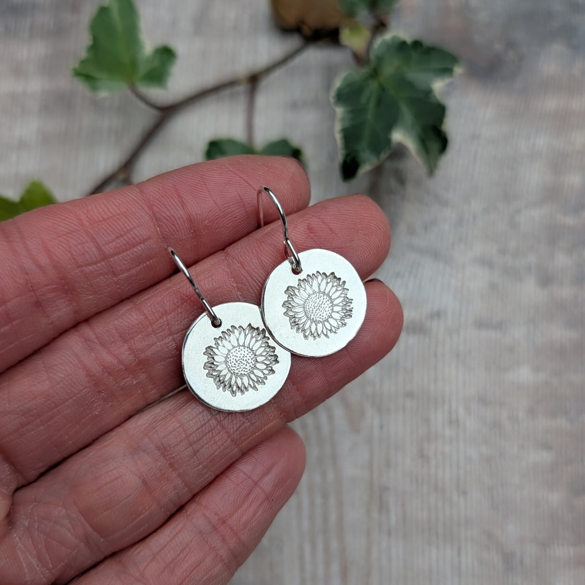 Handmade silver sunflower disc earrings held in a hand to show their size, featuring polished recycled sterling silver discs with stamped sunflower patterns and silver hook ear wires.