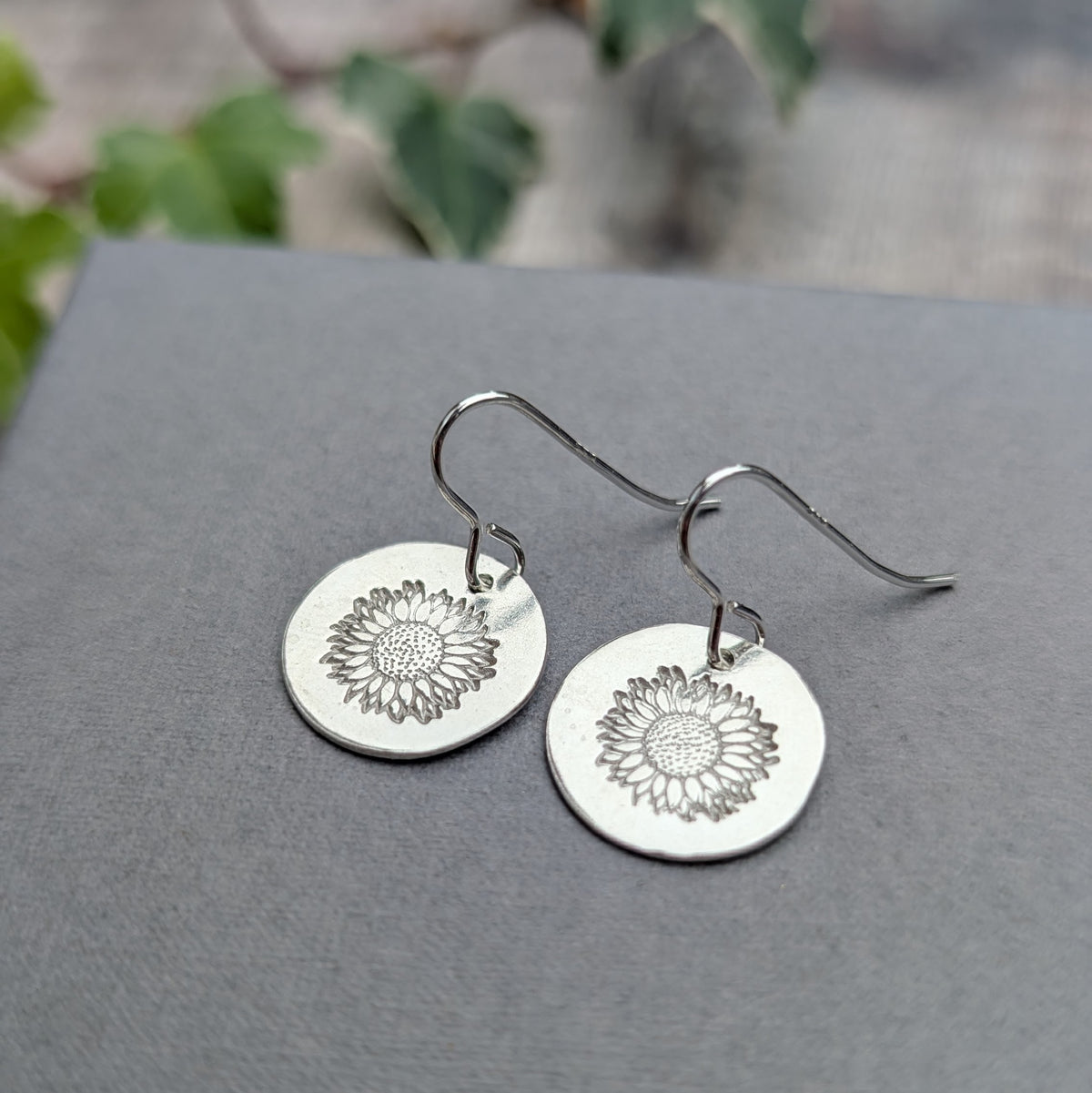 Silver sunflower disc earrings handmade from recycled sterling silver, featuring stamped sunflower designs on round silver discs with hook ear wires, displayed on a grey background.