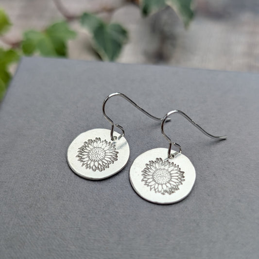 Silver sunflower disc earrings handmade from recycled sterling silver, featuring stamped sunflower designs on round silver discs with hook ear wires, displayed on a grey background.