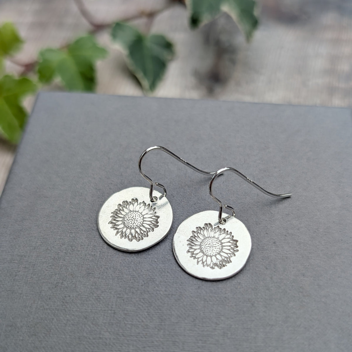 Handmade silver sunflower disc earrings with detailed sunflower patterns stamped into polished sterling silver discs, photographed on a grey surface with greenery in the background.