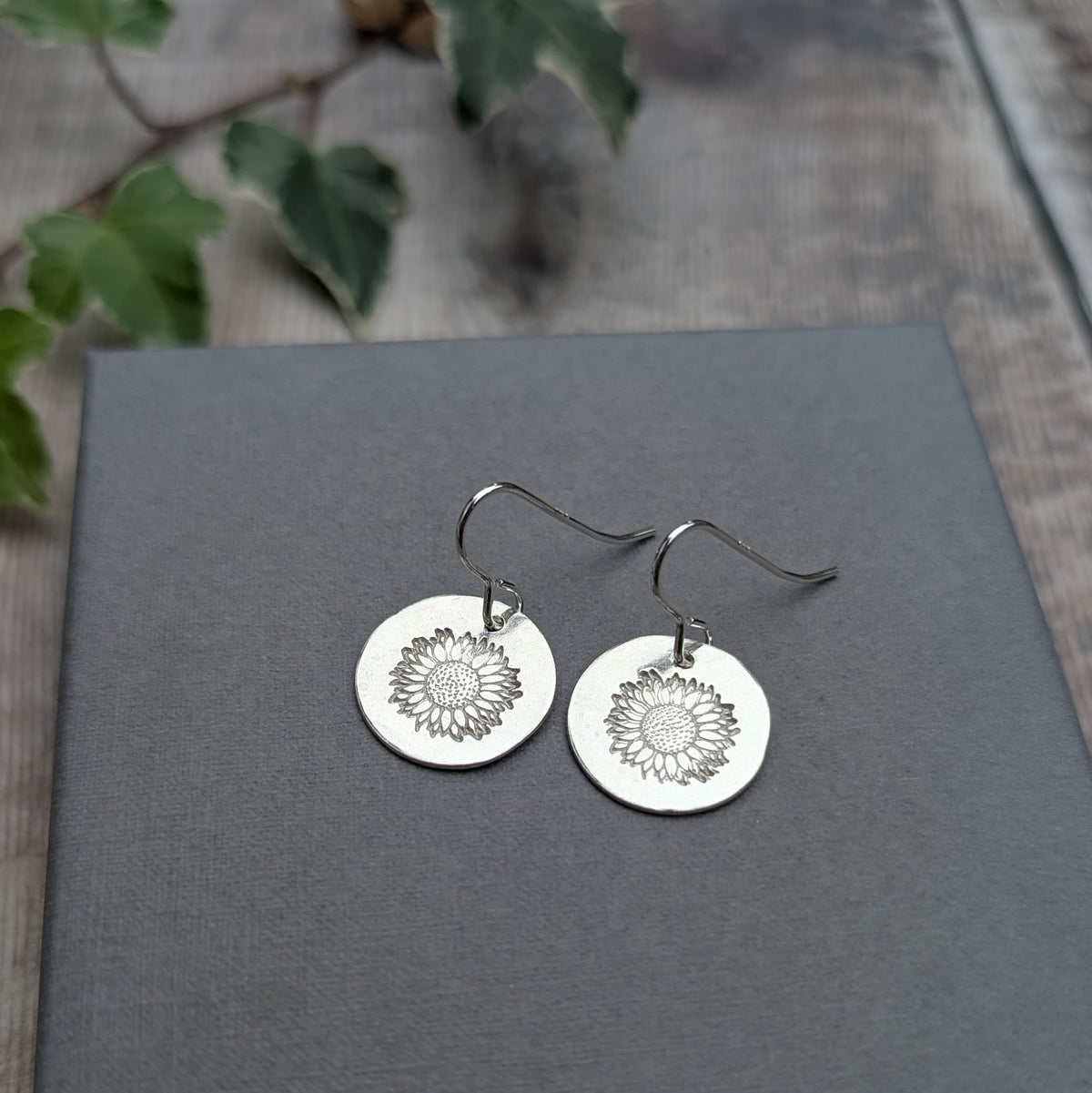 Pair of silver sunflower disc earrings made from recycled sterling silver, featuring round discs with stamped sunflower designs and silver hook ear wires, displayed on a grey background.